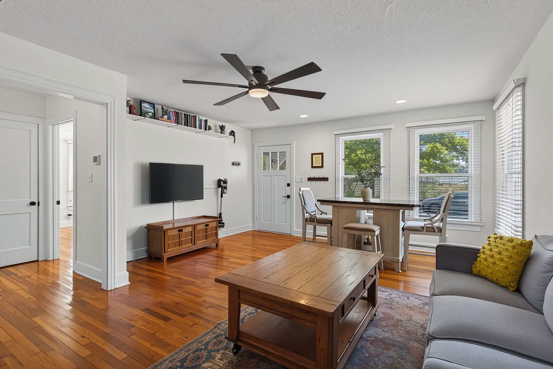 A spacious living room with hardwood flooring, a ceiling fan, a flat-screen TV, a wooden coffee table, a gray sofa with a yellow cushion, and a dining area with a wooden table and chairs.
