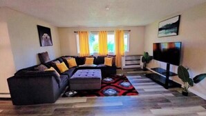 A modern living room with a sectional sofa, a flat-screen TV, and a large window with curtains. - Midnight Sun Viewpoint Aurora (Fairbanks)