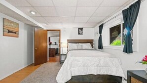 A bedroom with a bed, a nightstand, a window with curtains, a door leading to another room, and a small rug on the floor. - Alaska Rafiki - Unit 3 (Fairbanks)