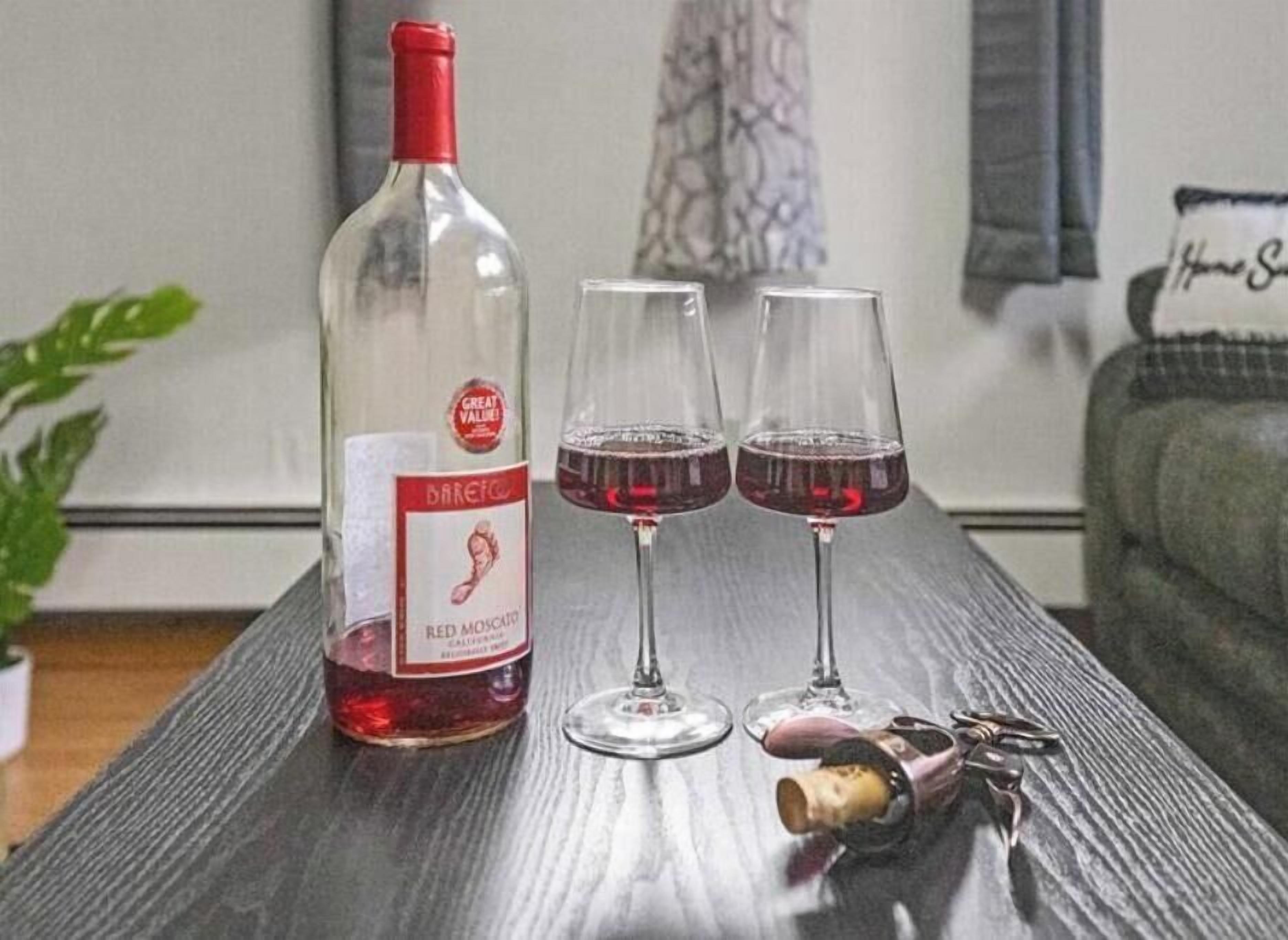 Pullo Barefoot Red Moscato -punaviiniä puupöydällä, sen vieressä kaksi viinilasia ja korkinirrotin.