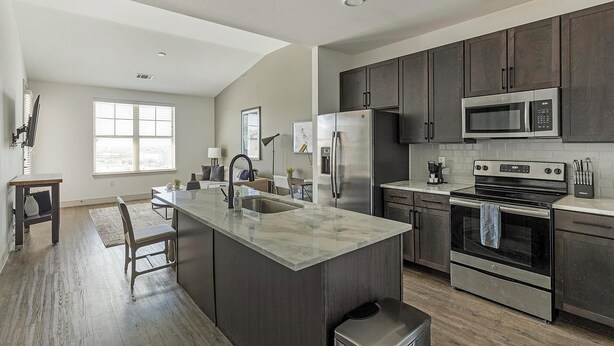 A modern kitchen with dark cabinets, stainless steel appliances, a marble island, and a large window providing natural light. - Landing | Beautiful 1BD, Clubhouse, Courtyard (Denton)