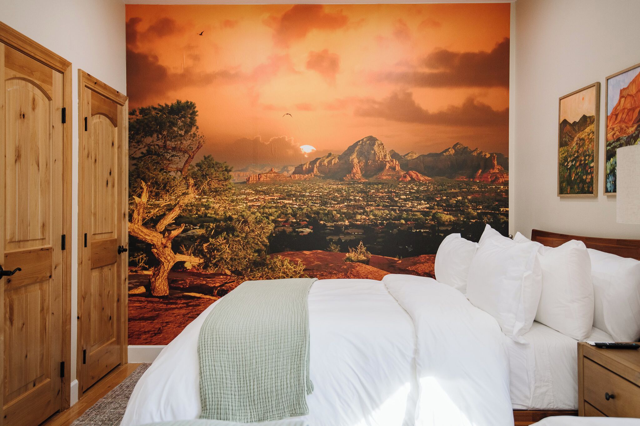 A bedroom with a large mural of a sunset landscape, a bed with white linens, and wooden doors.