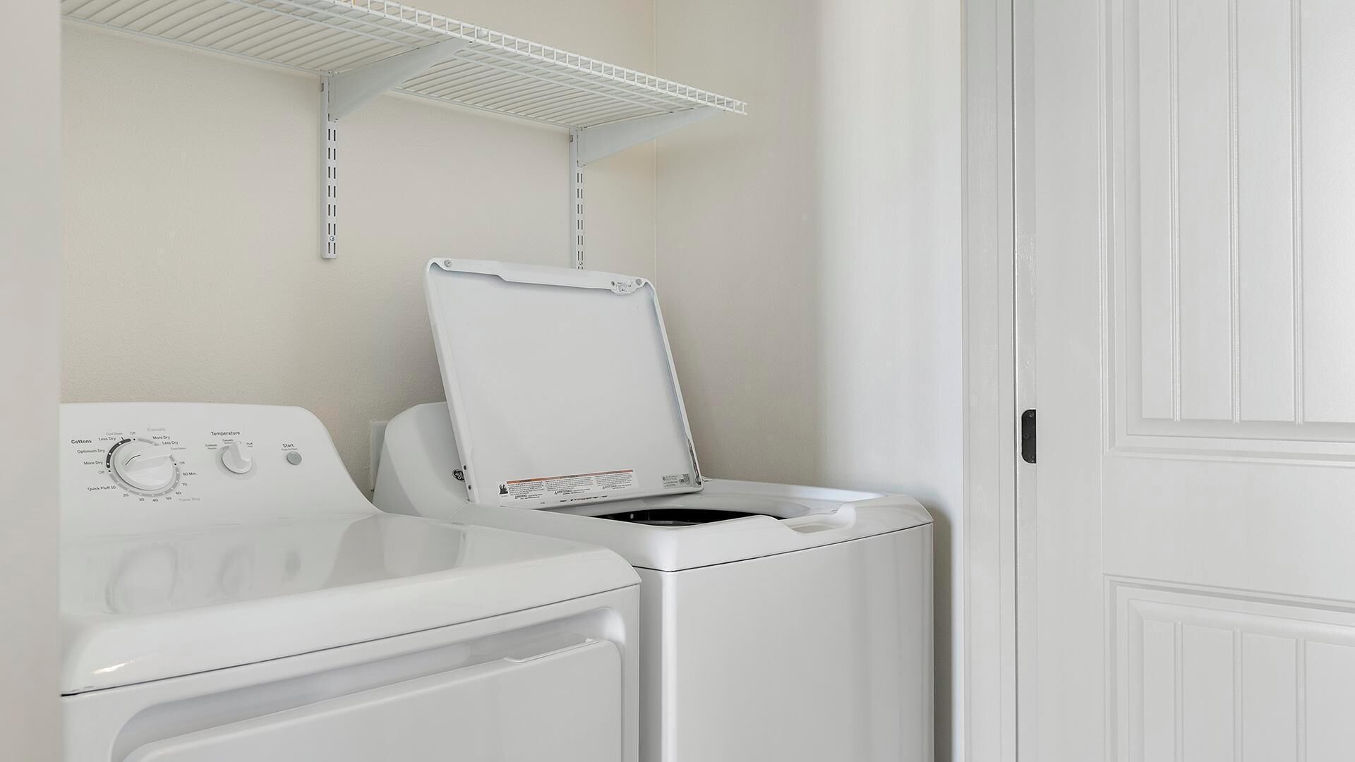 Une buanderie avec un lave-linge et un sèche-linge blancs, une étagère en métal au-dessus du sèche-linge et une porte blanche.