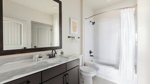 A bathroom with a large mirror, a bathtub with a shower curtain, a toilet, and a marble countertop. - Landing | Incredible 2BD, Clubhouse, Courtyard (Denton)
