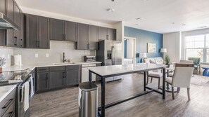 A modern kitchen with dark cabinets, a central island, and a dining area with a blue accent wall. - Landing | Modern 2BD, Clubhouse, Gym (Denton)