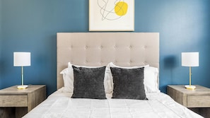 Un lit double avec une tête de lit capitonnée, deux tables de chevet avec des lampes et une œuvre d’art abstraite encadrée au-dessus du lit.