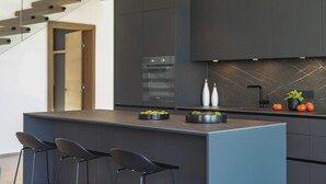 A modern kitchen with dark cabinetry, a central island with bar stools, a built-in oven, and a decorative backsplash. - Villa Moana on Krk - Crovillas (Vrbnik)