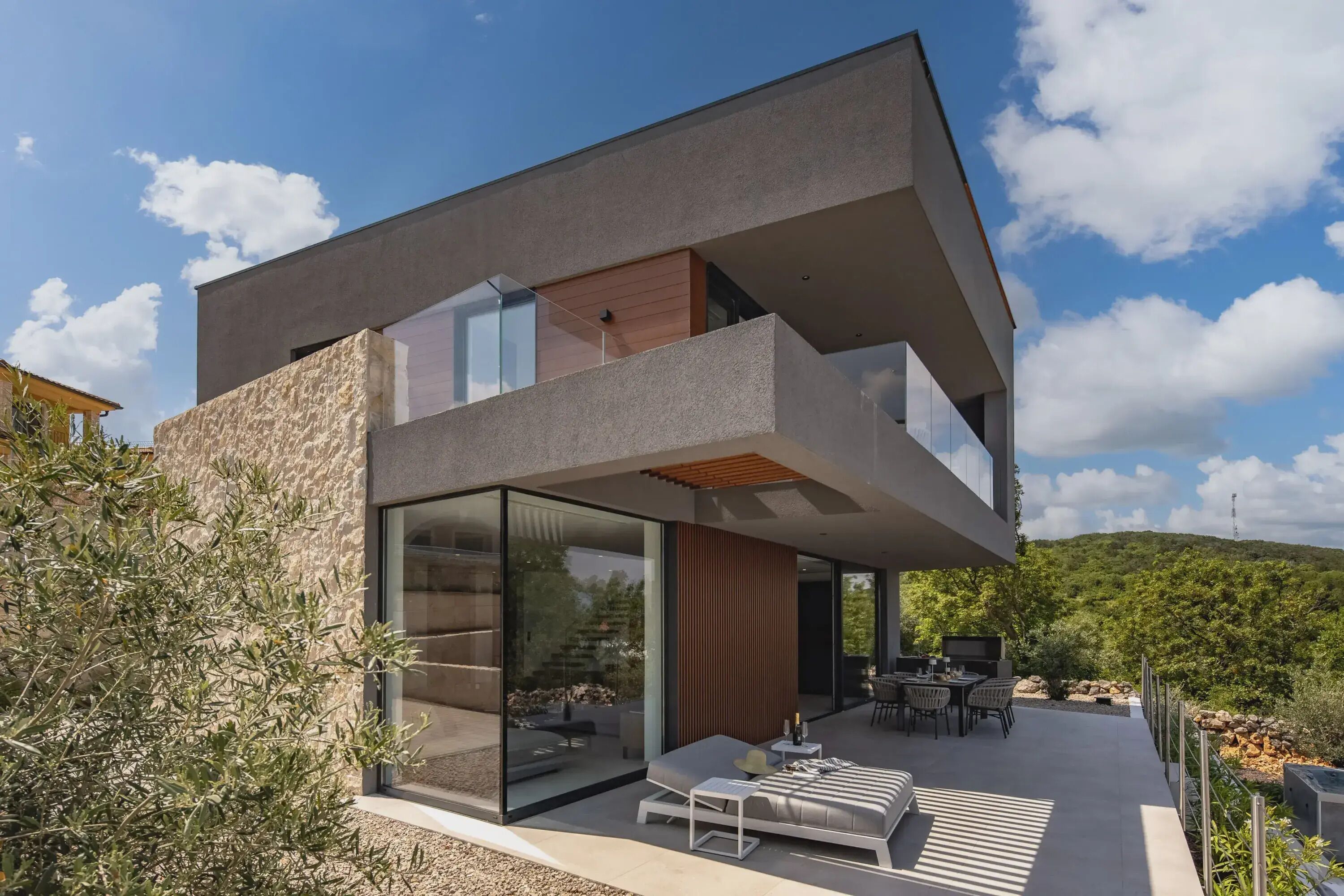 A modern two-story house with a balcony, outdoor seating area, and a view of the surrounding landscape.