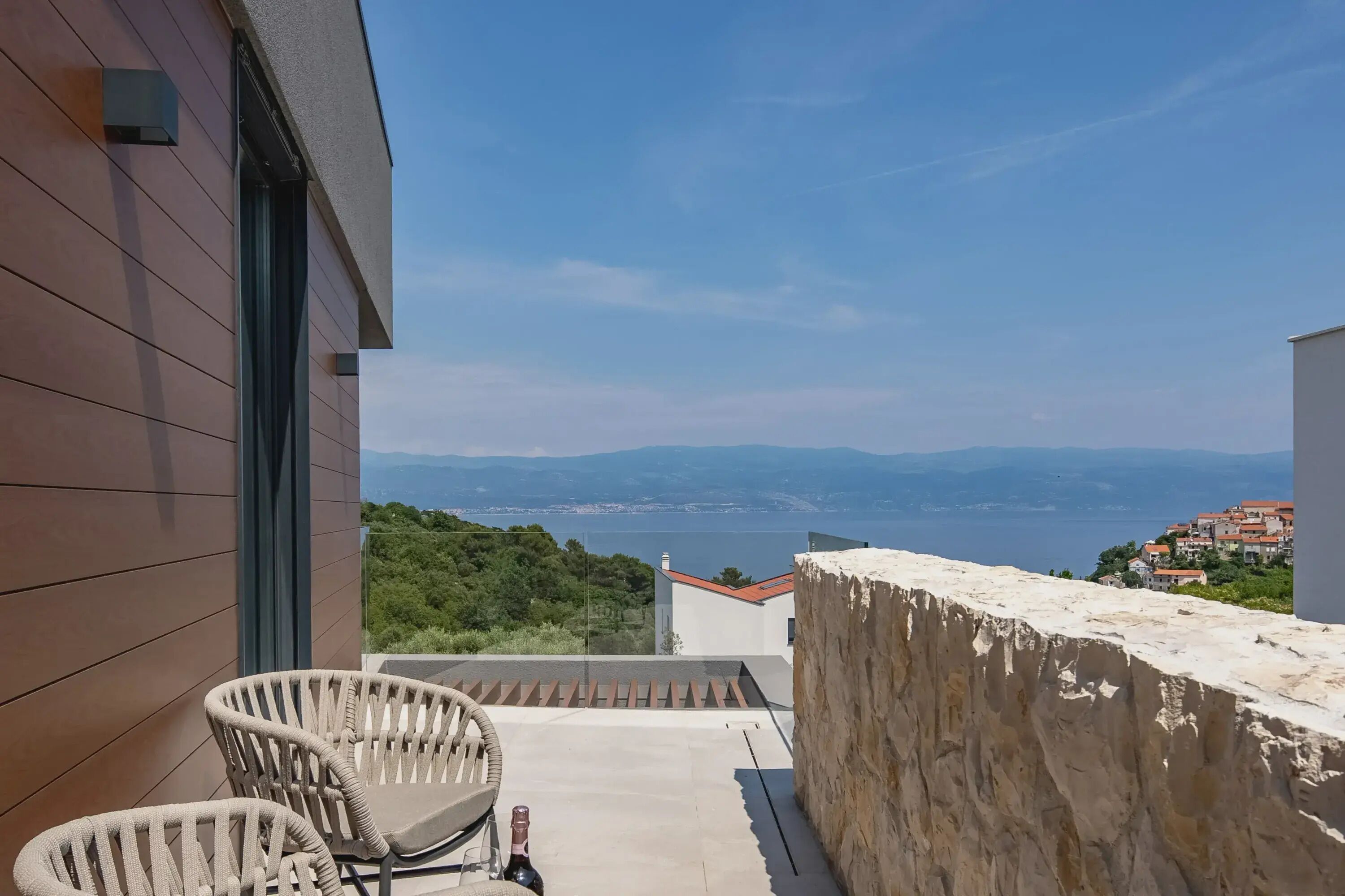 A balcony with two wicker chairs and a bottle of wine, overlooking a coastal landscape with buildings and mountains in the distance.