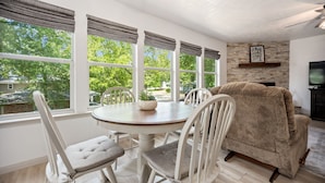 A dining area with a round table and four chairs, a sofa, and a television in a room with large windows and a stone wall. - New Listing - Cape Royale Tree House w/ Fire Pit – Direct Lake Access (Coldspring)