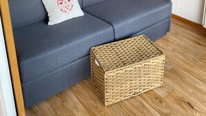 Huoneessa on harmaa sohva, jossa on koristeellinen koristetyyny, puinen kaappi ja rottinkikori. Seinät ovat vihreät ja lattia on puuta.