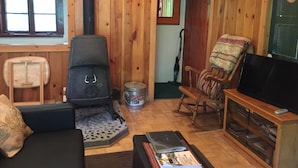 A cozy cabin interior with a wood-burning stove, a comfortable seating area, and a television on a wooden stand.