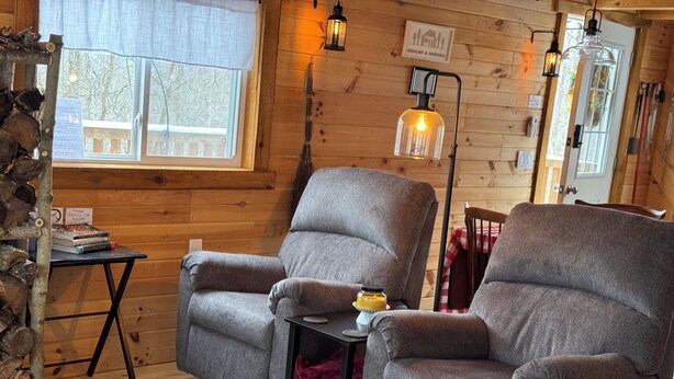 A cozy cabin interior with wooden walls, a large window, a fireplace, and a comfortable seating area with a table and a lamp.