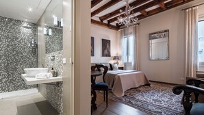 A modern bathroom with a mosaic tile wall, a white sink, and a shower. The bedroom features a large bed, a wooden floor, and a chandelier.