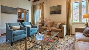 A living room with a blue armchair, a beige sofa, and a glass coffee table. There are paintings on the walls and a window with curtains.