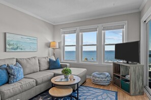 A living room with a sofa, a coffee table, a TV on a wooden stand, and a view of the sea through large windows.