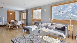 Een moderne woonkamer met een bank, een eettafel met stoelen en een groot raam met uitzicht op de bergen.