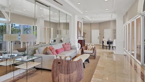 A spacious living area with a large sofa, a coffee table, and a wicker chair. There are glass tables with lamps, a dining area with chairs, and a view of the outdoors through large windows. - 2/2 Bay Front Condo, Pool, Private Beach Access (Longboat Key)