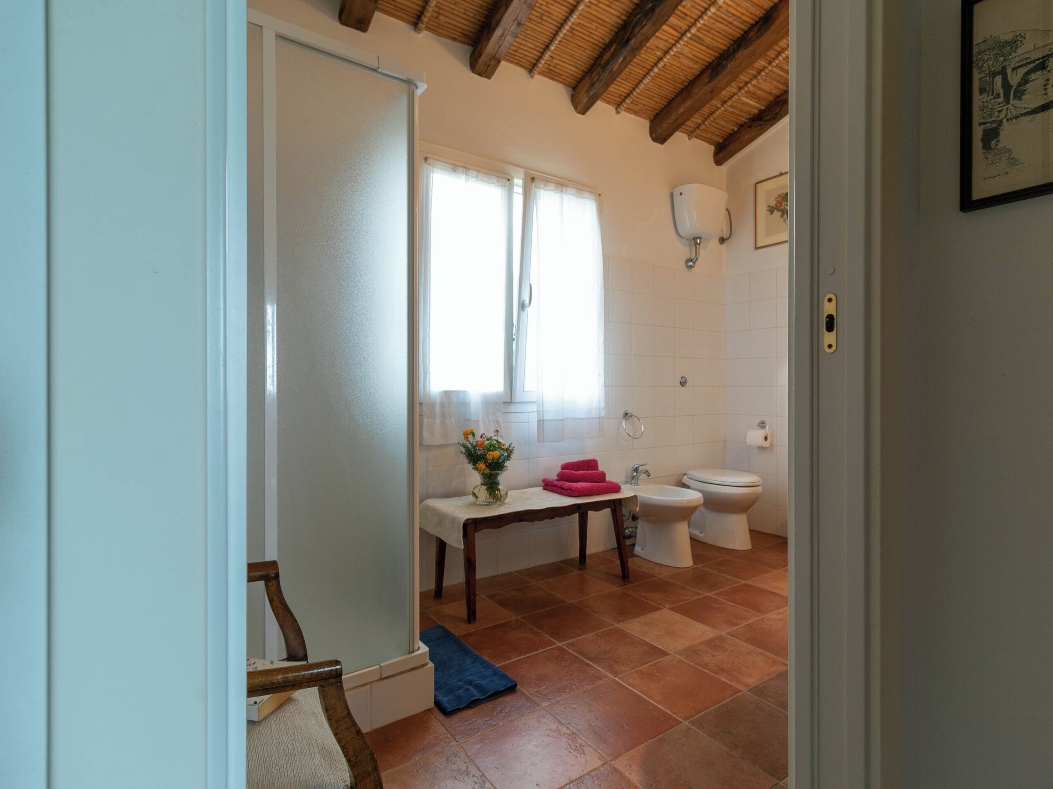 Un baño con inodoro, bidé y lavamanos. Hay una ventana con cortinas, un perchero y una mesita con un jarrón de flores.