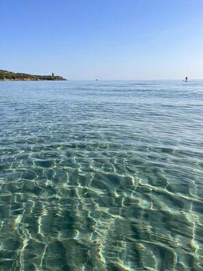 A clear view of a calm sea with gentle waves, a distant shoreline with a tower, and a person paddleboarding in the distance. - Villa Clara in Tertenia (Tertenia)