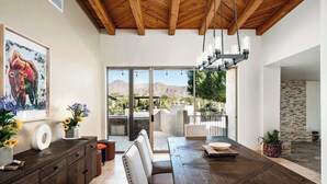 Uma sala de jantar com mesa de madeira, assentos acolchoados, lustre e vista para montanhas através de grandes portas de vidro.