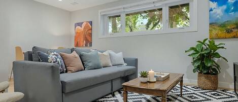 A modern living room with a grey sofa, patterned rug, and a large window with a view of greenery.