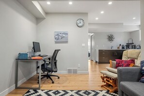 A modern living room with a desk, a comfortable sofa, a mounted clock, and a kitchen area in the background. - Cozy and comfy two-unit home in the Calgary core (Calgary)