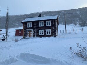 A wooden cabin with a snow-covered roof, a wooden door, and a small porch area. - Old log cabin from 1830 with soul. (Trøndelag)