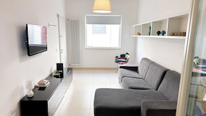 Ett modernt vardagsrum med en grå soffa, en platt-TV som är monterad på väggen, ett svart sidobord med en lampa och böcker, och ett fönster med utsikt över omgivningen.