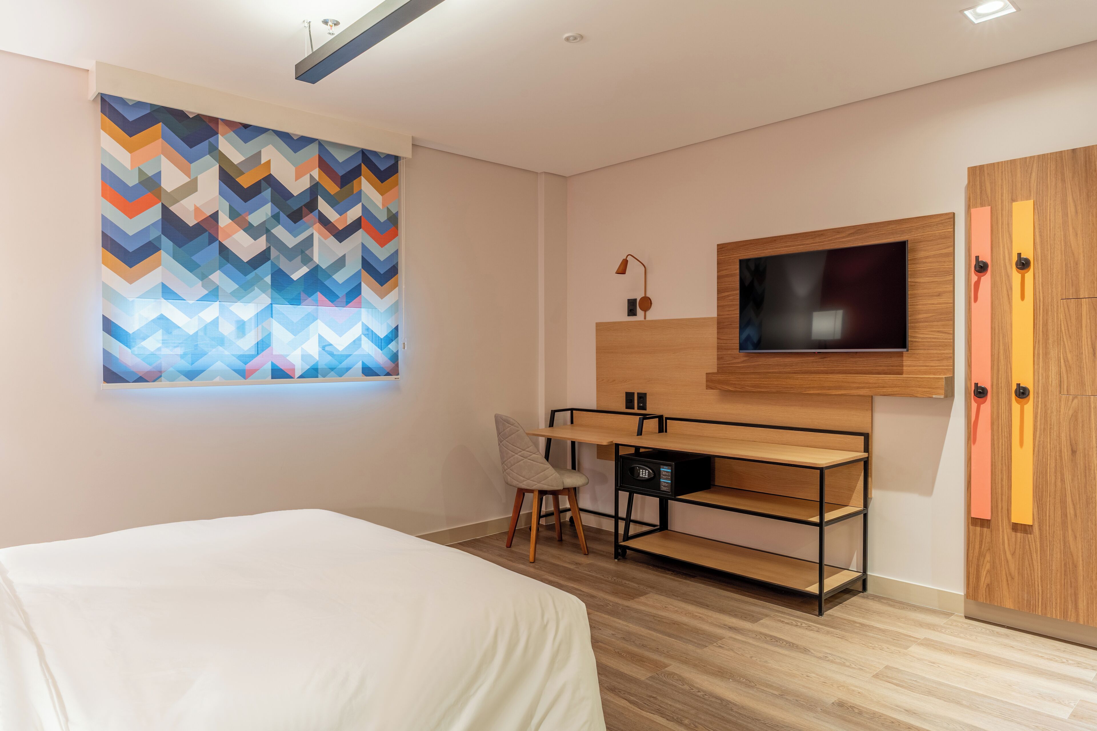 Une chambre d’hôtel moderne équipée d’un lit, d’un bureau avec une chaise, d’une télévision et d’une œuvre d’art murale aux couleurs vives.