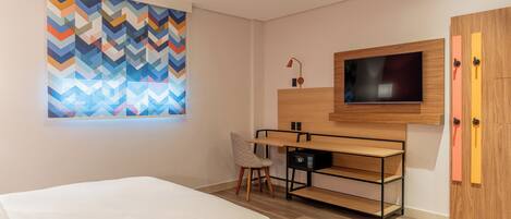 Quarto de hotel moderno com uma cama, uma escrivaninha com cadeira, uma televisão e uma obra de arte colorida na parede.
