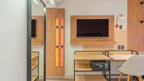A modern hotel room with a wooden wardrobe, a flat-screen TV mounted on the wall, a desk with a chair, and a bedside table with a lamp.
