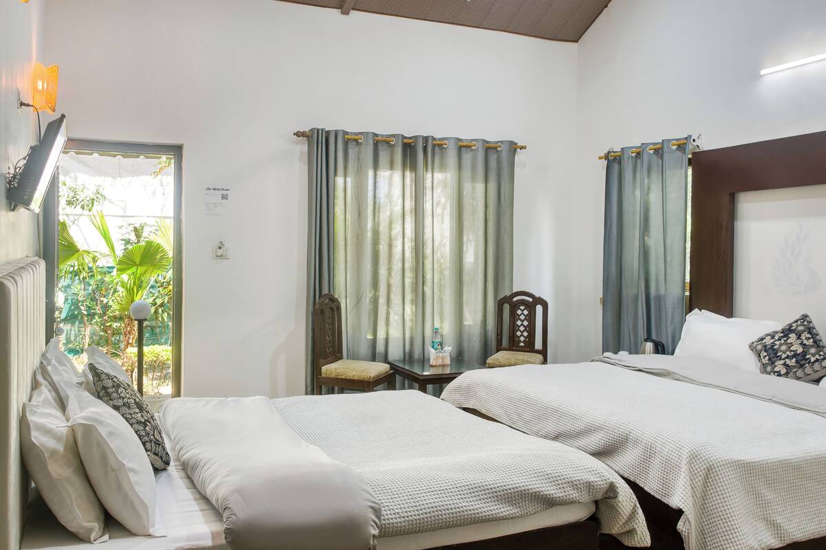 A hotel room with two beds, a small table with a bottle, a chair, and a view of greenery outside.
