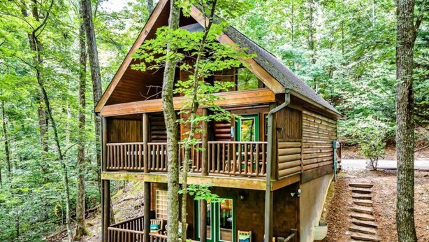 Una cabaña de madera de dos pisos con balcón, rodeada de bosque.