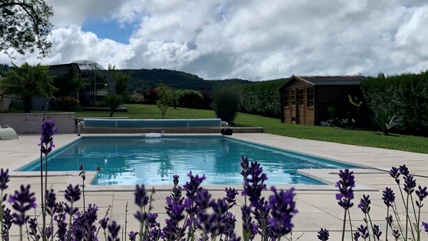 A rectangular swimming pool with a clear blue water surface, surrounded by a paved deck. In the background, there is a grassy area with a wooden cabin and a modern building with a glass facade.