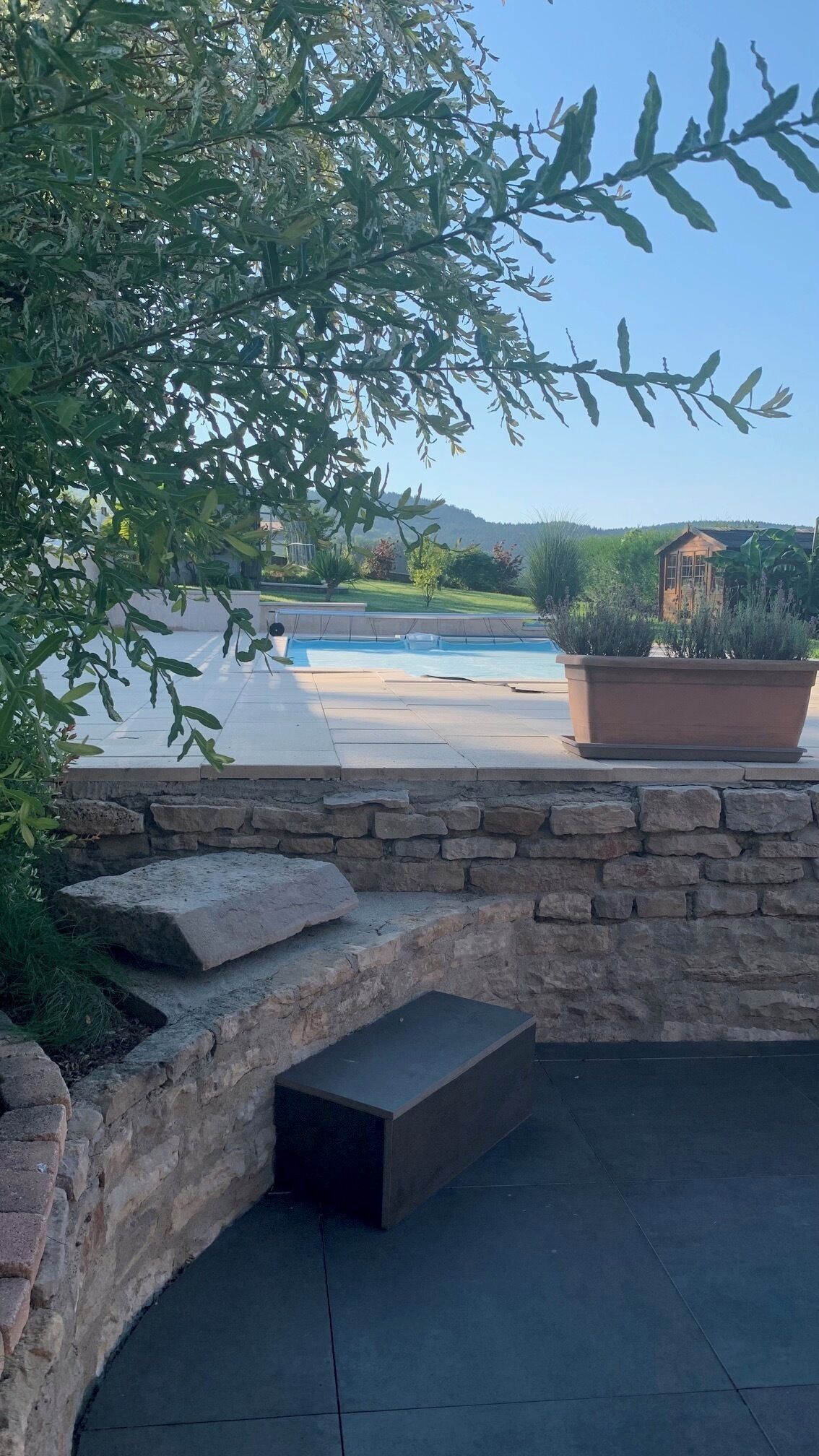 A stone patio with a pool, surrounded by greenery and a view of hills.