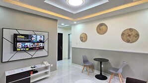 Un área de espera moderna con un televisor de pantalla plana montado en la pared, dos sillas grises, una mesita redonda y decoración mural.