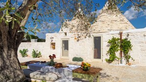 Un bâtiment en pierre surmonté d’un toit conique, un petit jardin avec une table et des coussins, et un arbre aux branches d’olivier.