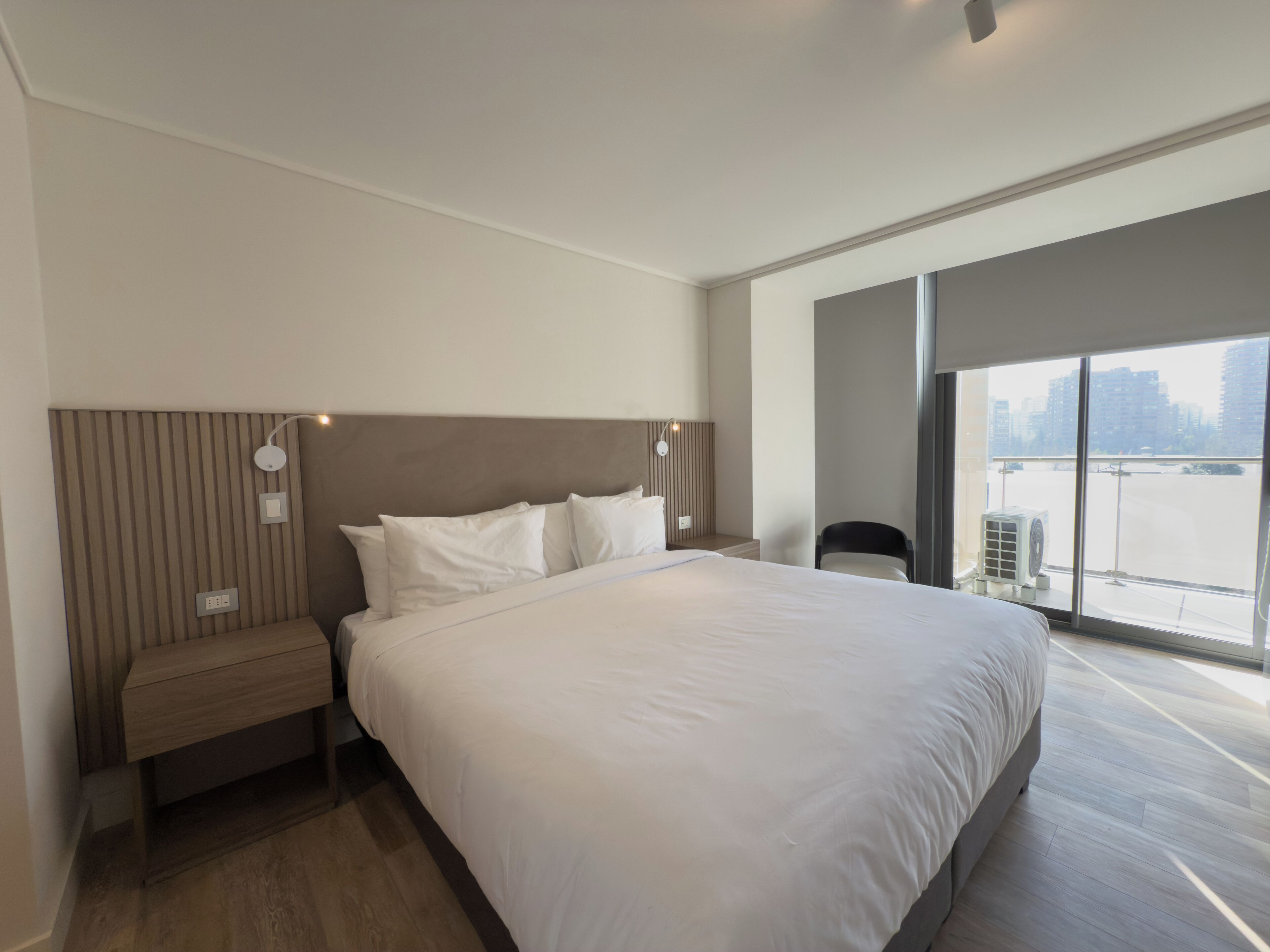 Una habitación de hotel moderna con una cama grande, una mesita de noche, una silla y vistas a la ciudad a través de amplios ventanales.