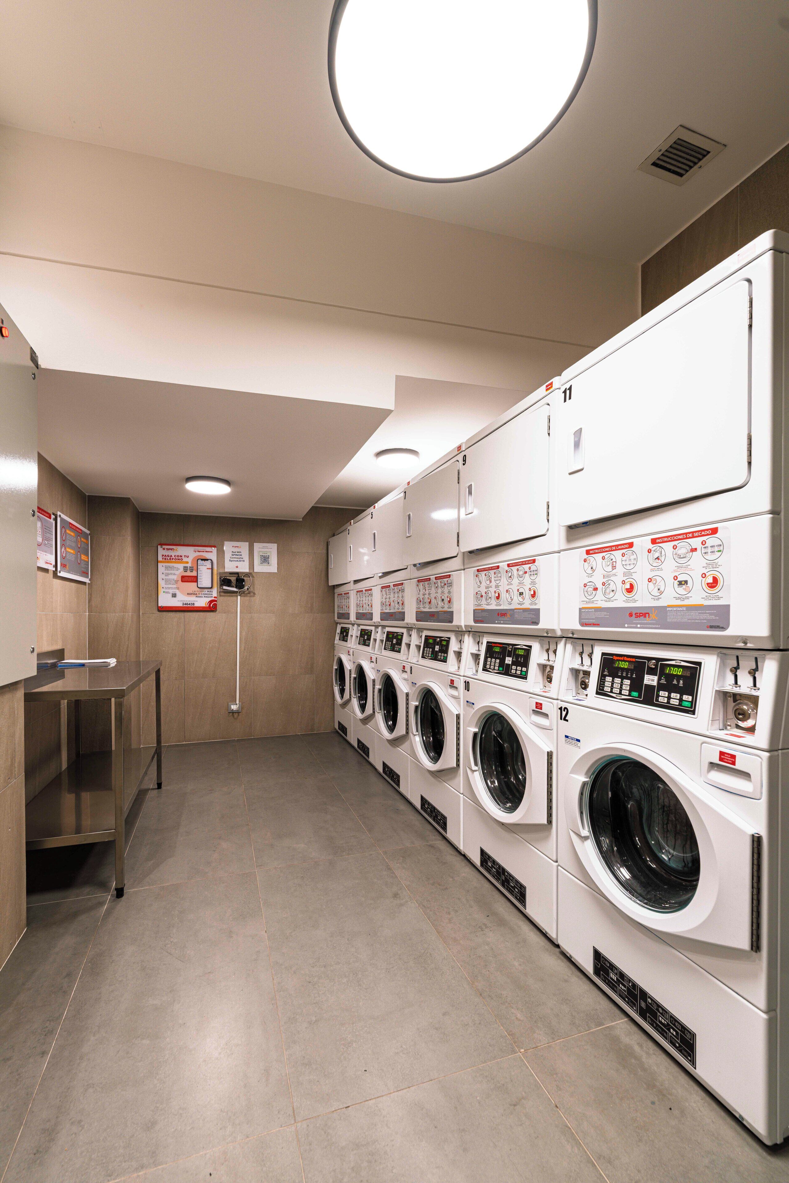 A laundry facility with multiple washing machines and dryers, a metal table, and informational signs on the wall.