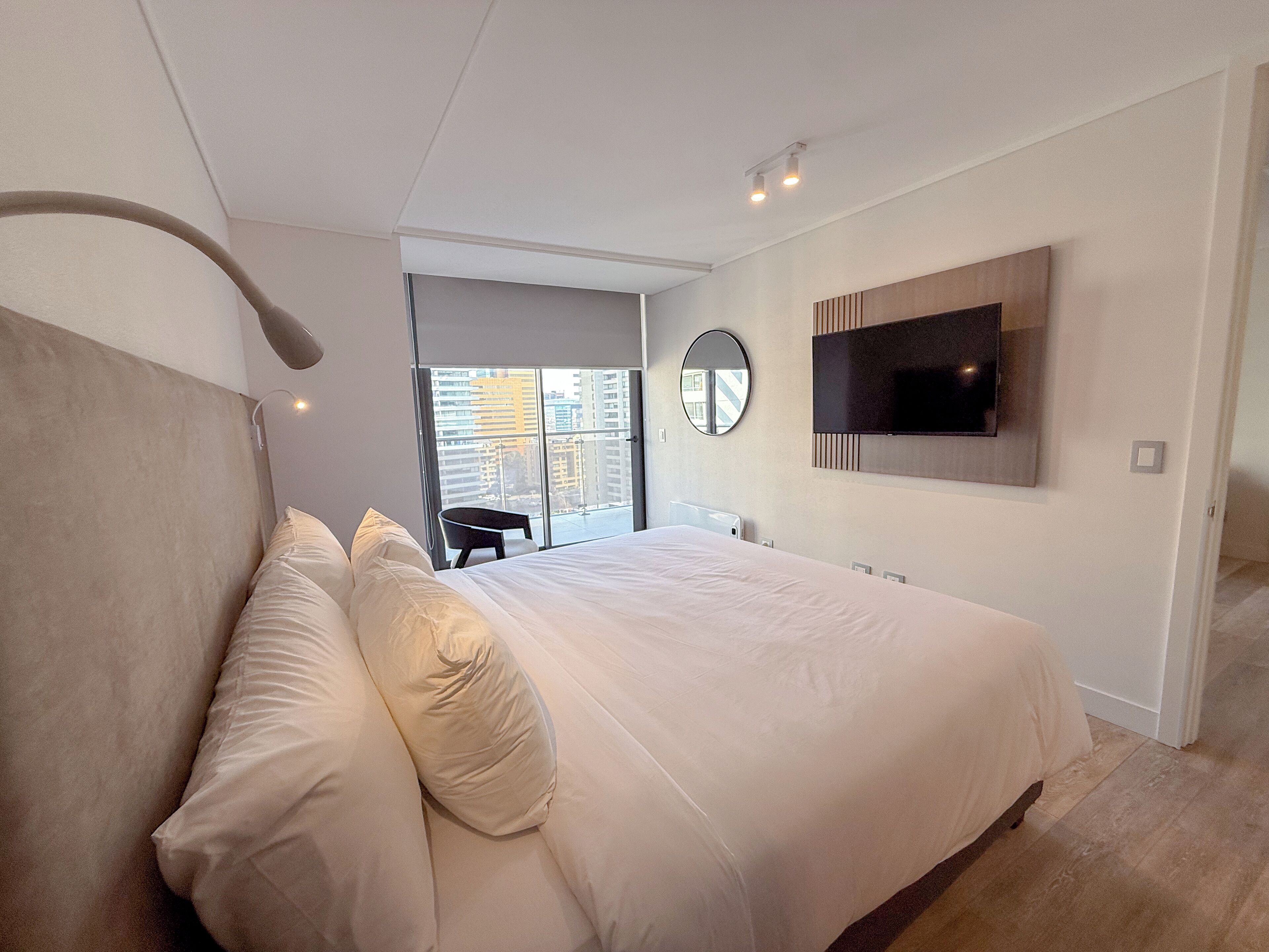 A modern bedroom with a large bed, a TV mounted on the wall, and a view of the city through the window.
