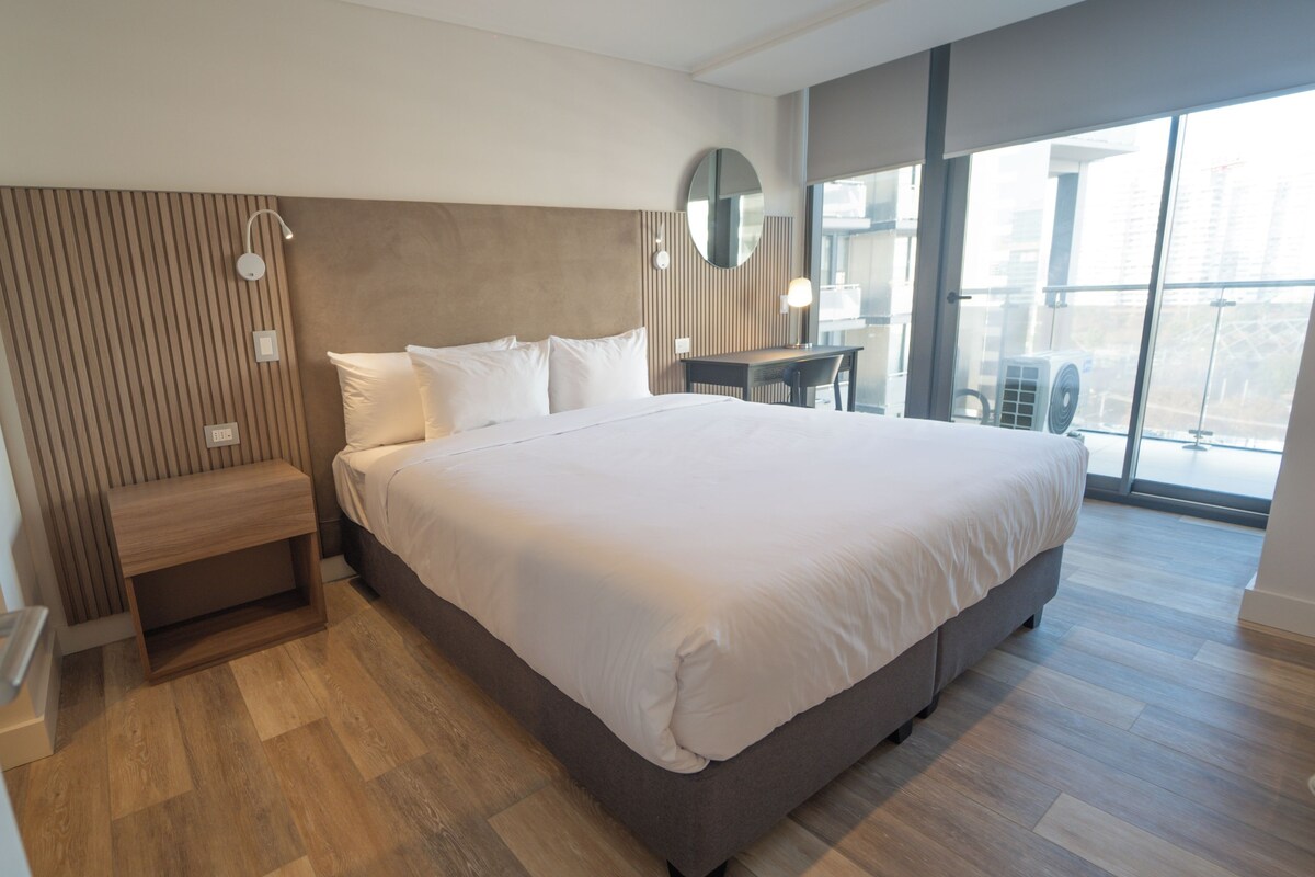 A modern hotel room with a large bed, a wooden headboard, a bedside table, a mirror, and a large window offering a city view.