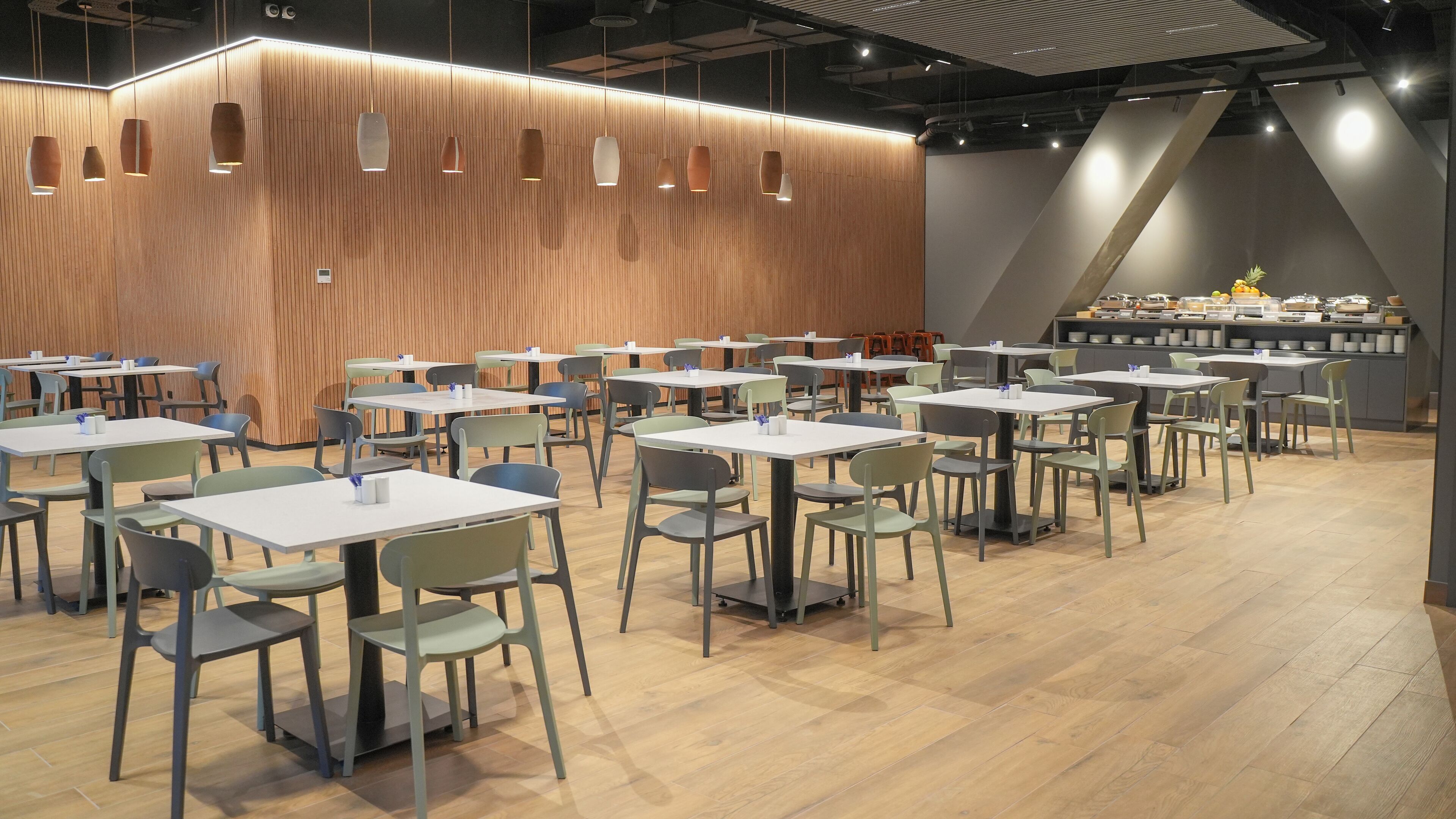 A modern dining area with wooden panel walls, multiple tables and chairs, and a food display counter.