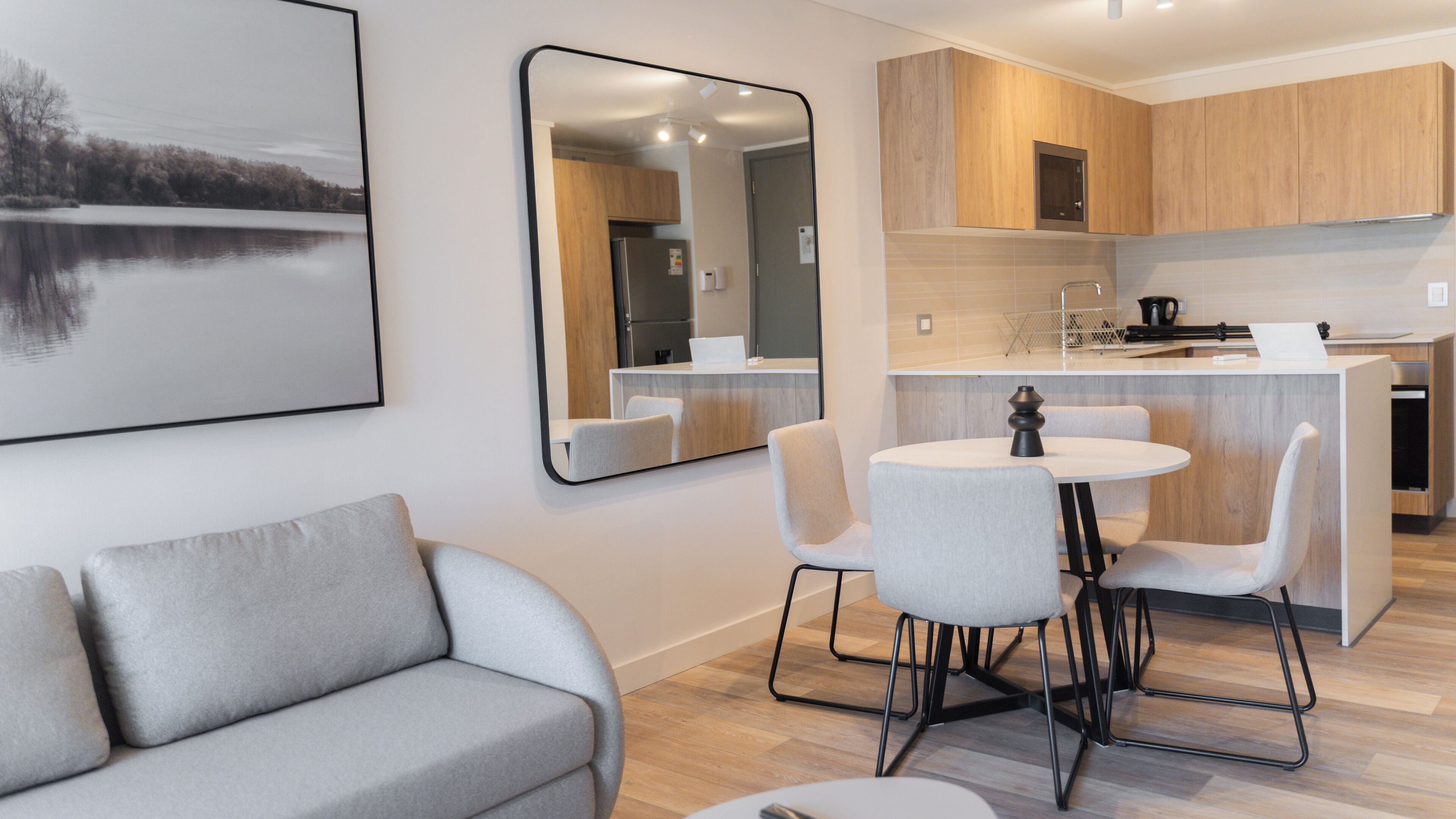 A modern living room with a grey sofa, a dining area with a round table and chairs, and a kitchen with wooden cabinets and a stainless steel refrigerator.