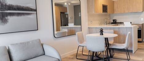 A modern living room with a grey sofa, a dining area with a round table and chairs, and a kitchen with wooden cabinets and a stainless steel refrigerator.