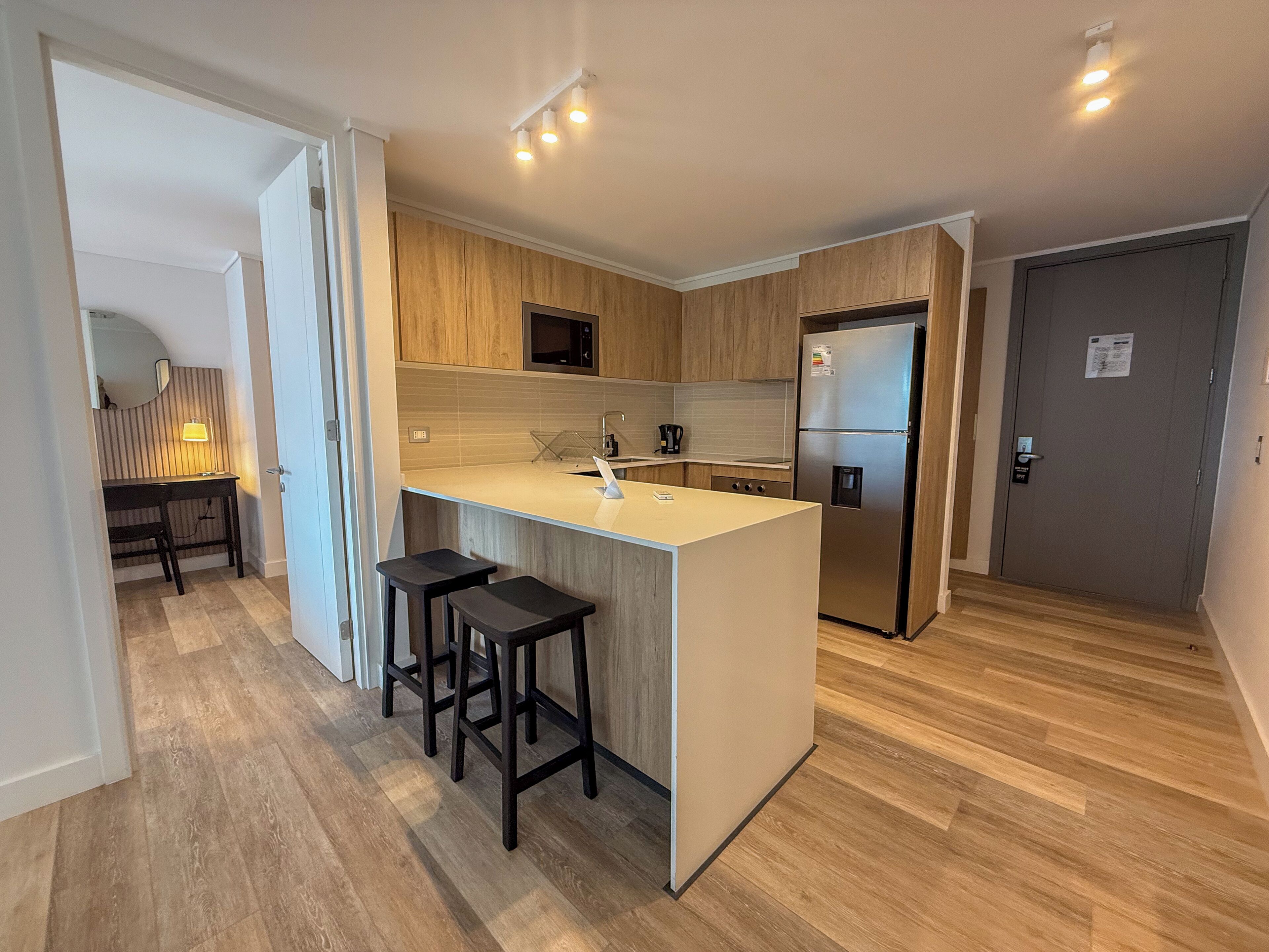 Une cuisine moderne avec des meubles en bois, un réfrigérateur en acier inoxydable, un plan de travail blanc et deux tabourets de bar.