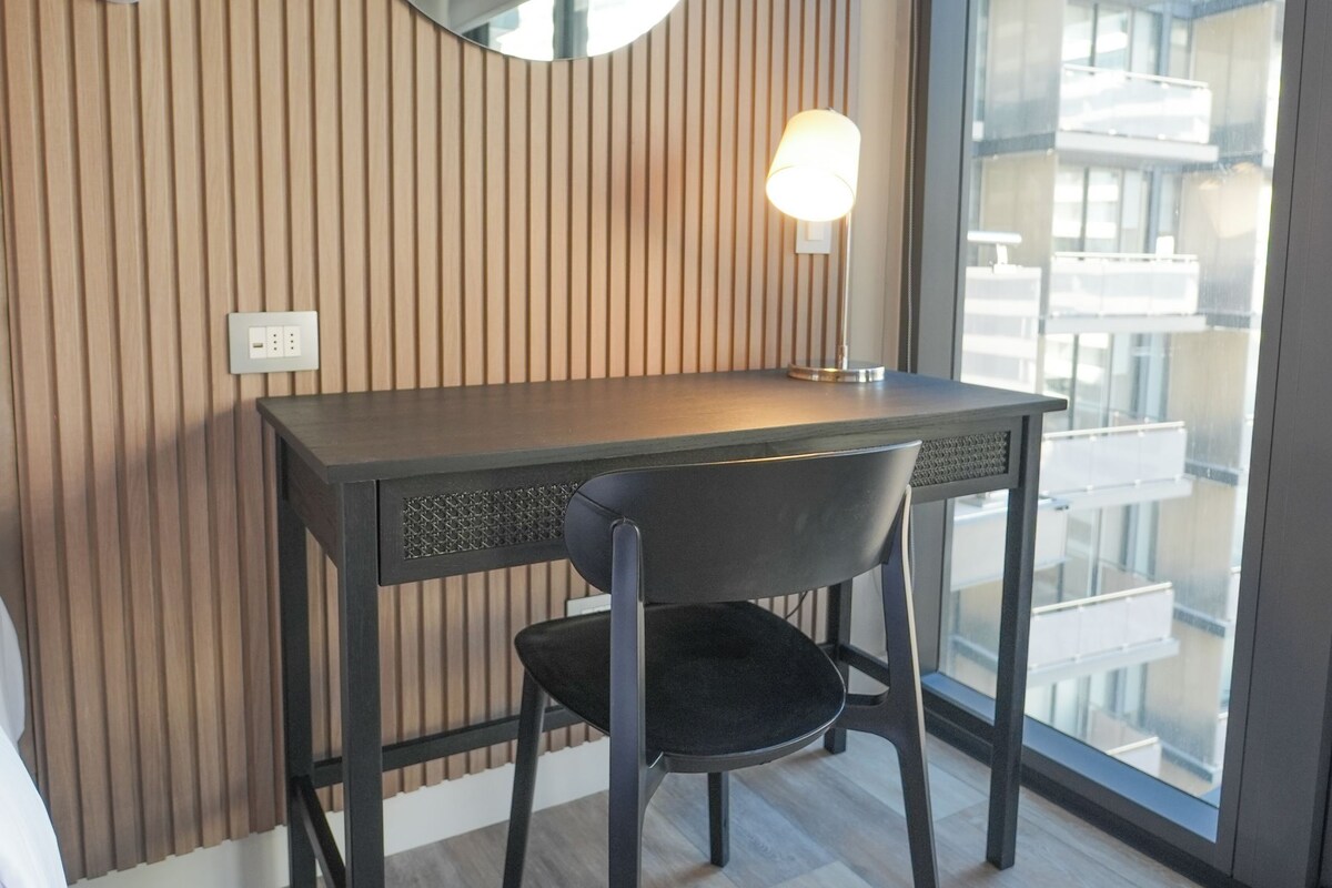 A modern room with a wooden slatted wall, a round mirror, a desk with a chair, and a large window offering a view of buildings.