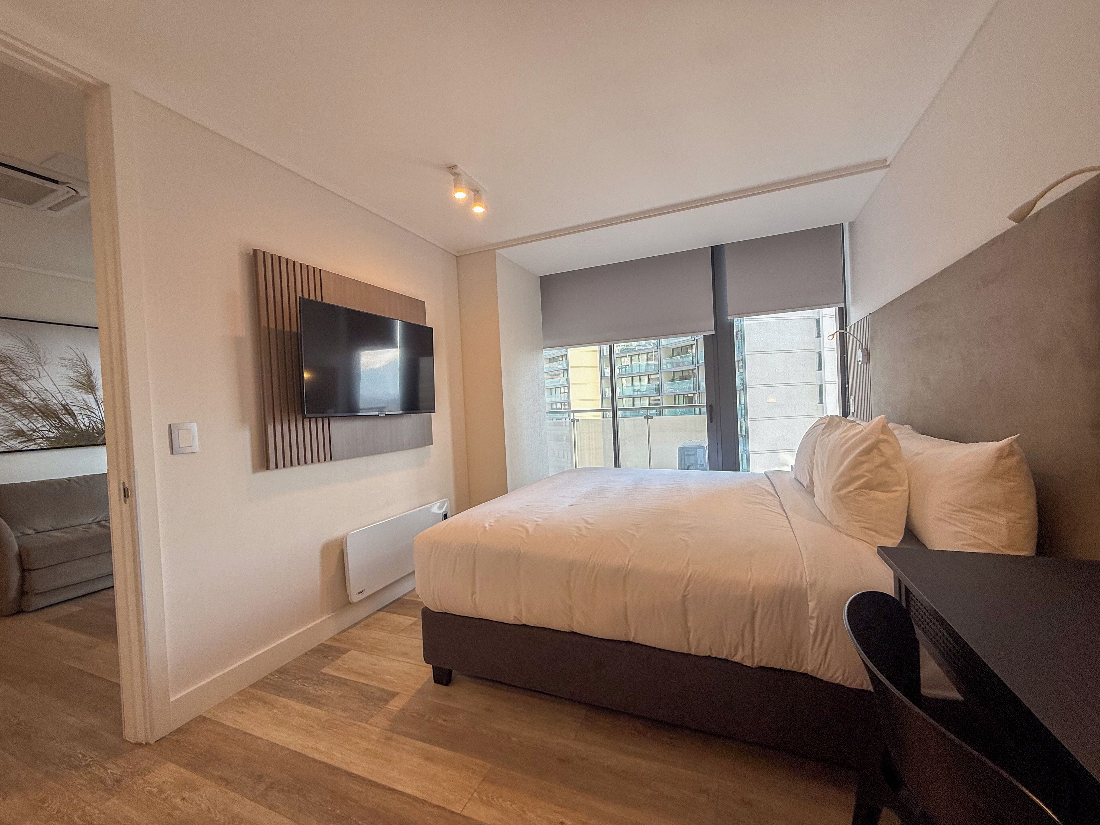A modern bedroom with a large bed, a flat-screen TV mounted on the wall, and a view of the city through the window.