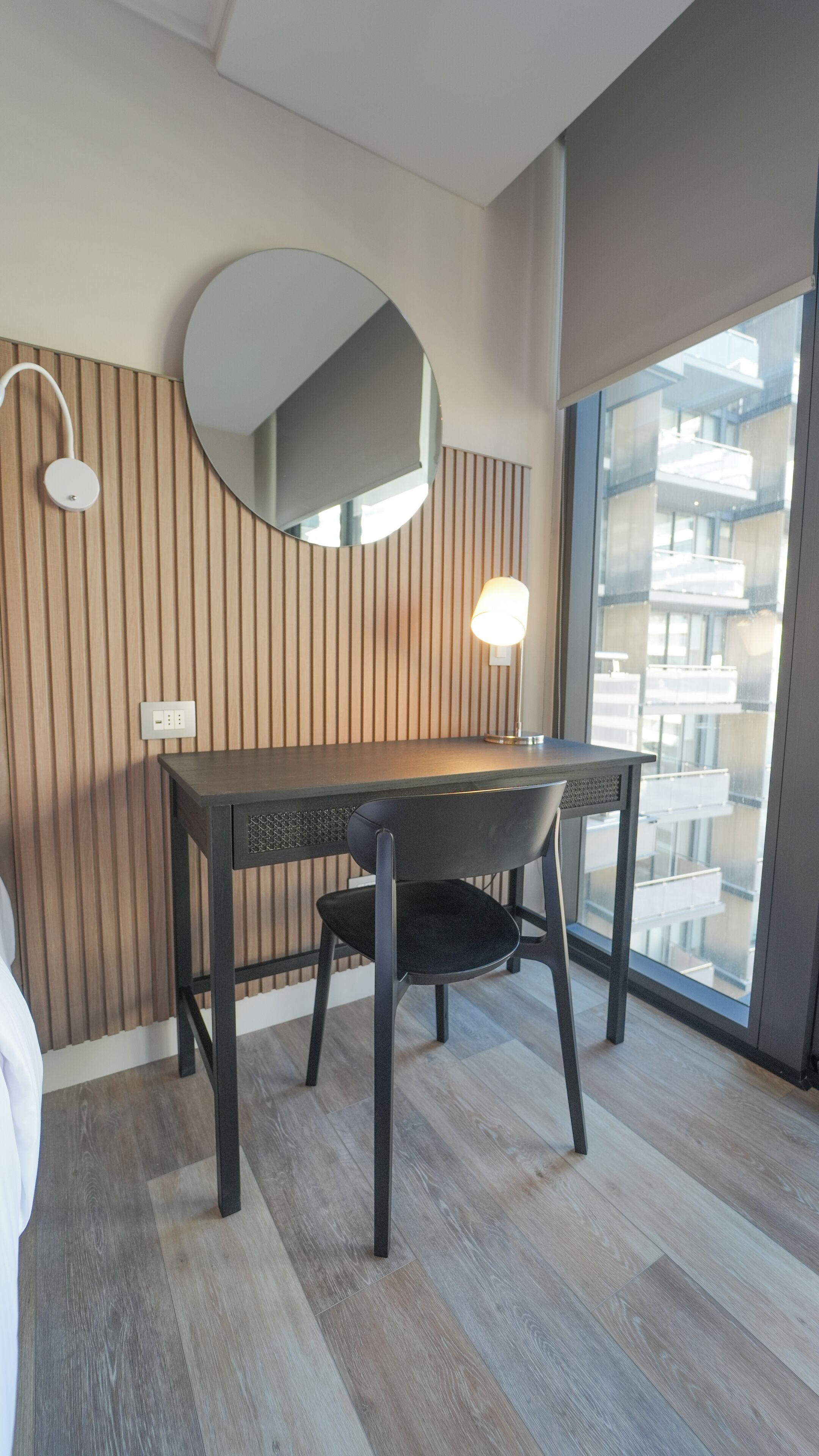 A modern room with a wooden slatted wall, a round mirror, a desk with a chair, and a large window offering a view of buildings.