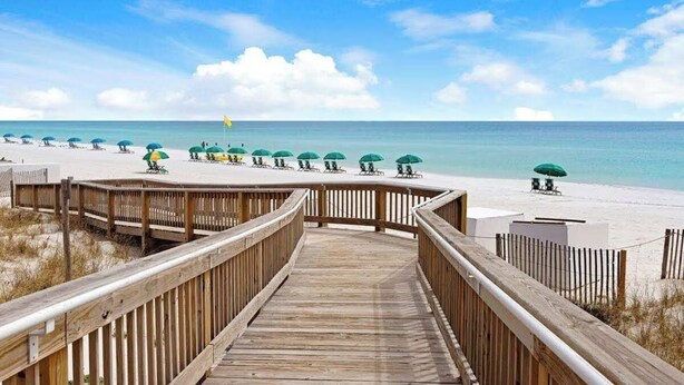 Une promenade en bois mène à une plage où s’alignent des rangées de parasols verts et de chaises longues.